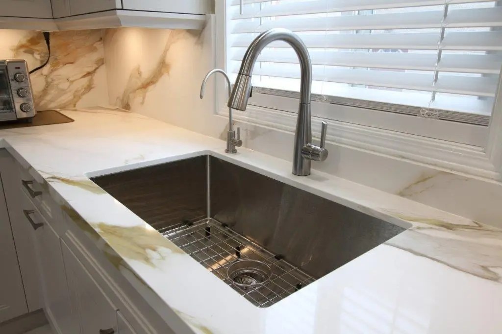 Modern kitchen with a stainless steel sink and chrome faucet, set in a sleek porcelain countertop with a matching backsplash; a toaster is visible on the counter, and a window with blinds is above the sink.