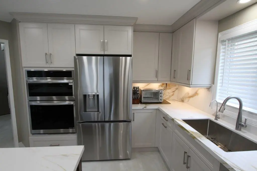 A modern kitchen with sleek white cabinetry, a stainless steel double-door refrigerator, built-in ovens, a countertop toaster oven, and a double basin sink under a window with blinds.