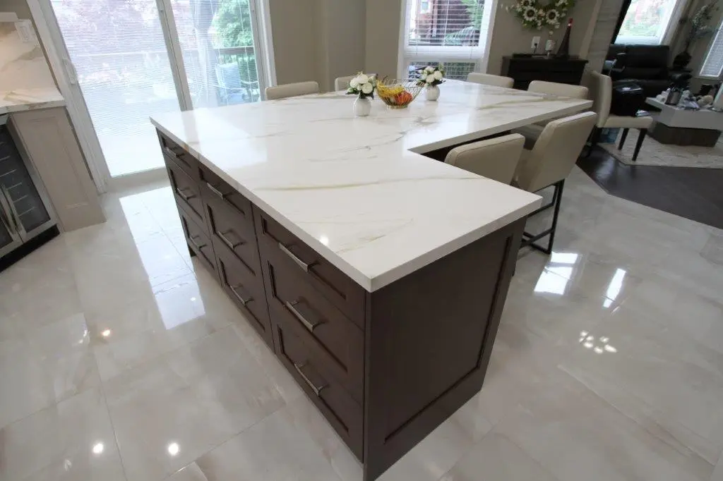 A modern kitchen island with a quartz top and wooden base featuring multiple drawers, surrounded by beige chairs on a polished tile floor.