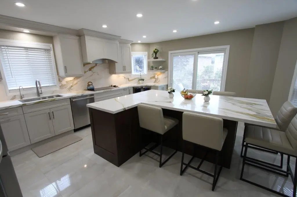 A modern kitchen with white cabinetry, porcelain countertops, and a central island with beige cushioned chairs. The kitchen features stainless steel appliances, including a dishwasher, and large windows allowing natural light to illuminate the space.