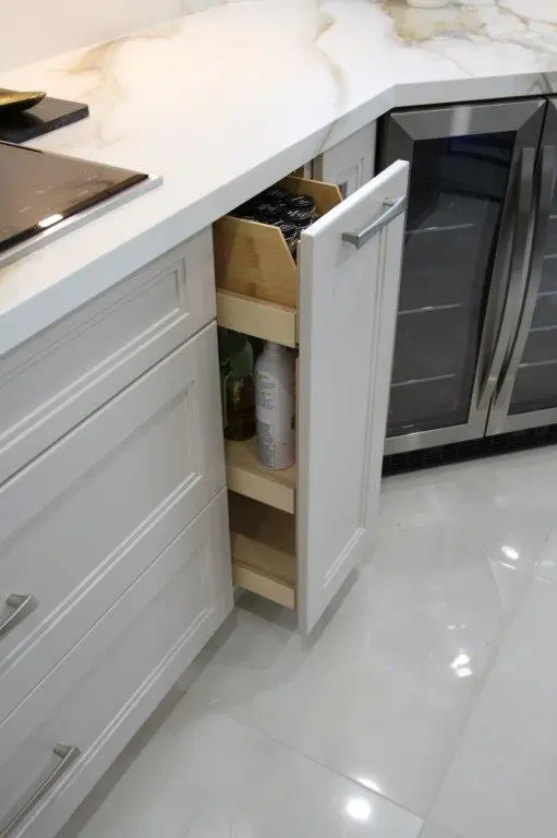 A kitchen with white cabinetry featuring a slim pull-out pantry, next to a section of porcelain countertop and a built-in stainless steel fridge.