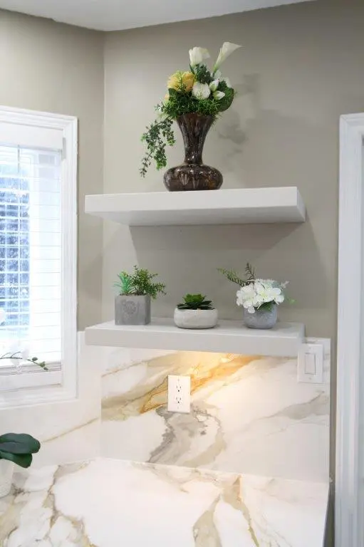 A corner of a kitchen counter featuring two floating shelves with decorative plants and flowers in vases, situated above a porcelain-patterned backsplash and countertop. A nearby window offers natural light to the scene.