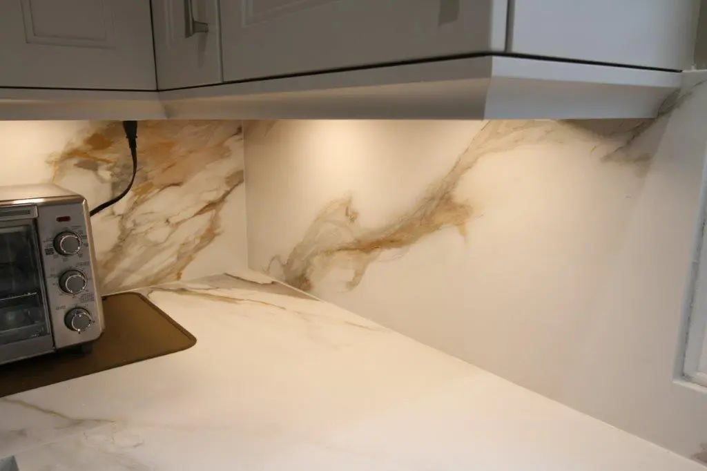 A kitchen countertop with a porcelain surface and backsplash, featuring warm beige and brown veining. Under-cabinet lighting illuminates the space.