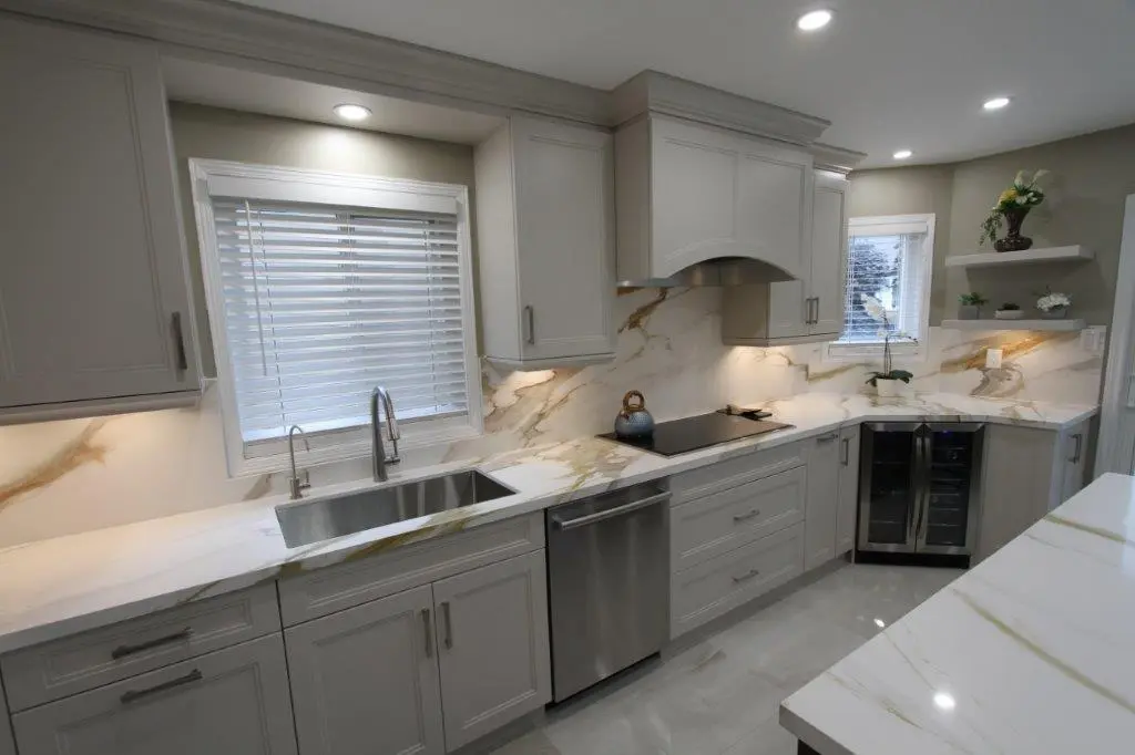 A modern kitchen with light gray cabinets, porcelain countertops and backsplash, a stainless steel double sink, built-in appliances, and recessed lighting.