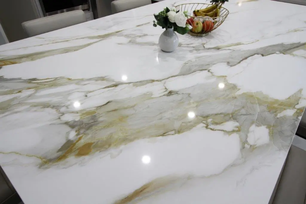 A polished porcelain countertop with white and gold veining, featuring a vase with white flowers and a fruit bowl containing bananas and apples in the background.