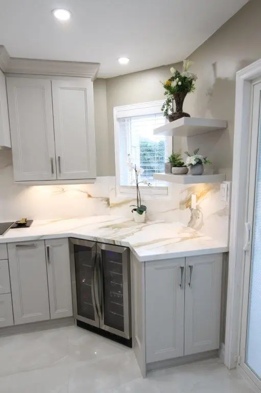 Modern kitchen corner featuring light gray cabinets and a Porcelain countertop, with a small wine fridge, plants on floating shelves, and a window with blinds.