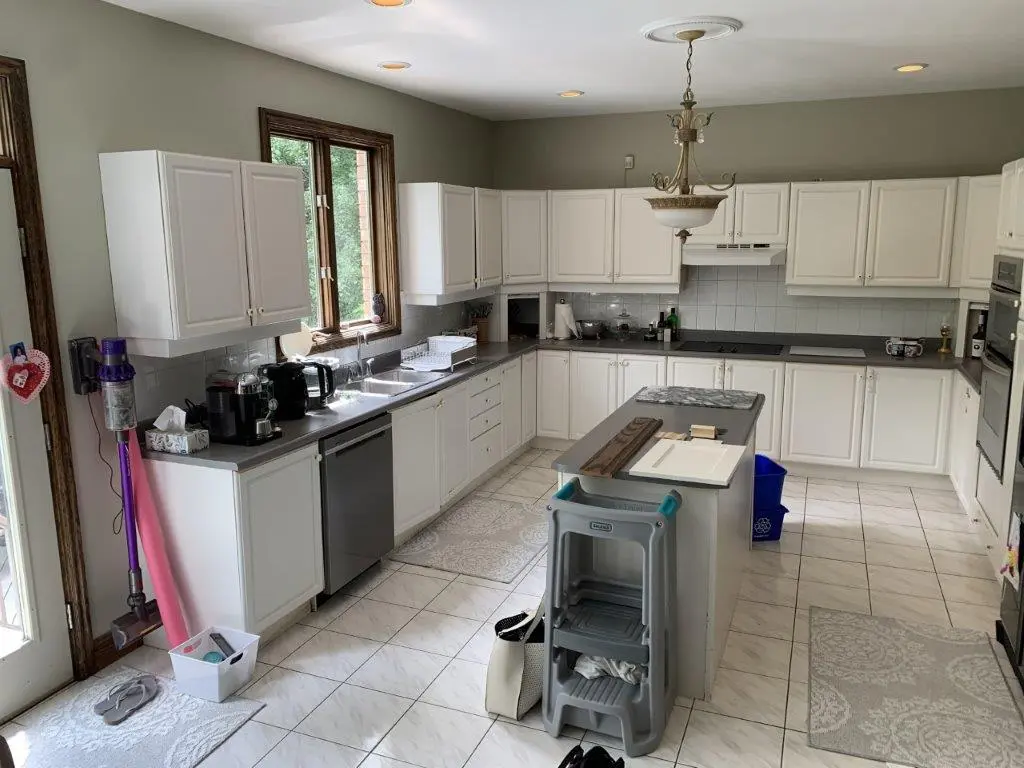 A before renovation kitchen with white cabinets, a black countertop, and tiled flooring. A large window provides natural light. The room is equipped with a dishwasher, sink, and various small appliances on the countertop.