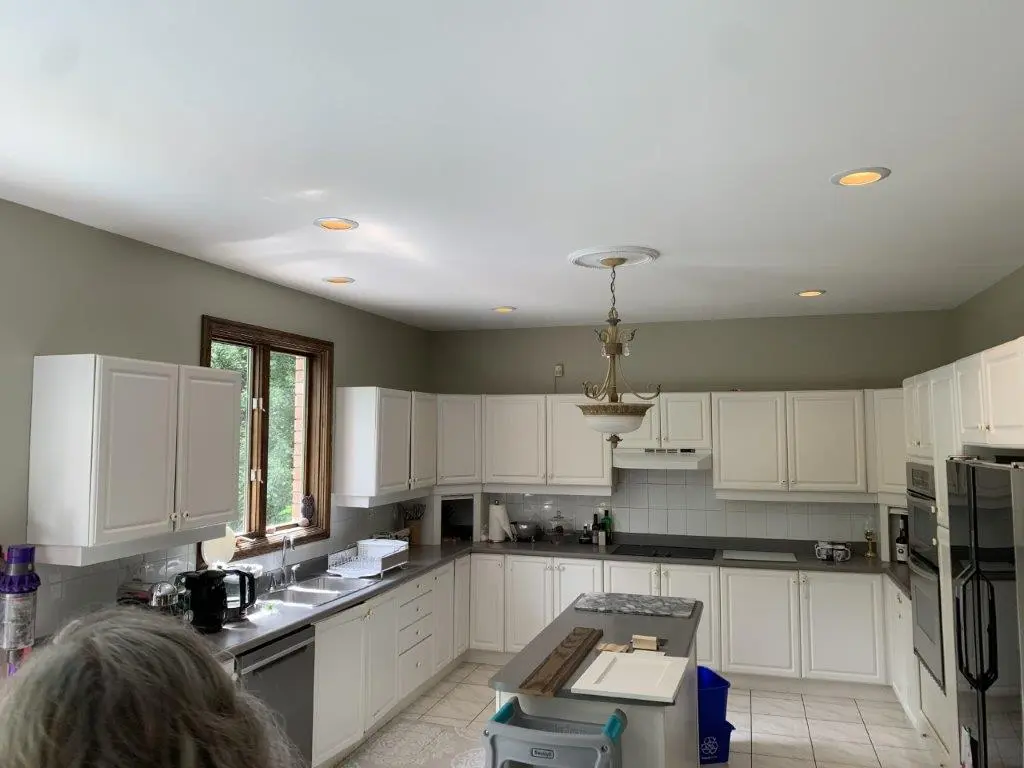 A before renovation kitchen with white cabinets and black countertops, centered around a small island with a wooden top. The room features a chandelier, multiple recessed lights, and a window to the left providing natural light. Appliances include a double oven and dishwasher, with various kitchen items on the counter.