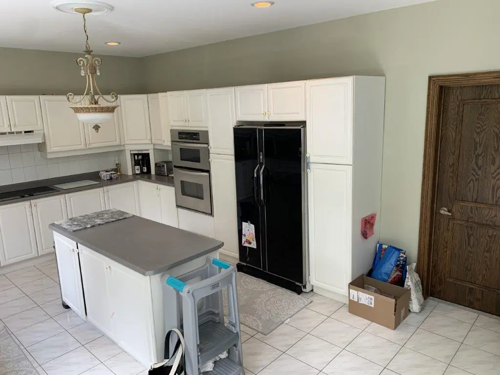 A before renovation kitchen with white cabinets, black appliances, and a central island with a grey countertop. The room features a small chandelier, tiled flooring, and a wooden door.