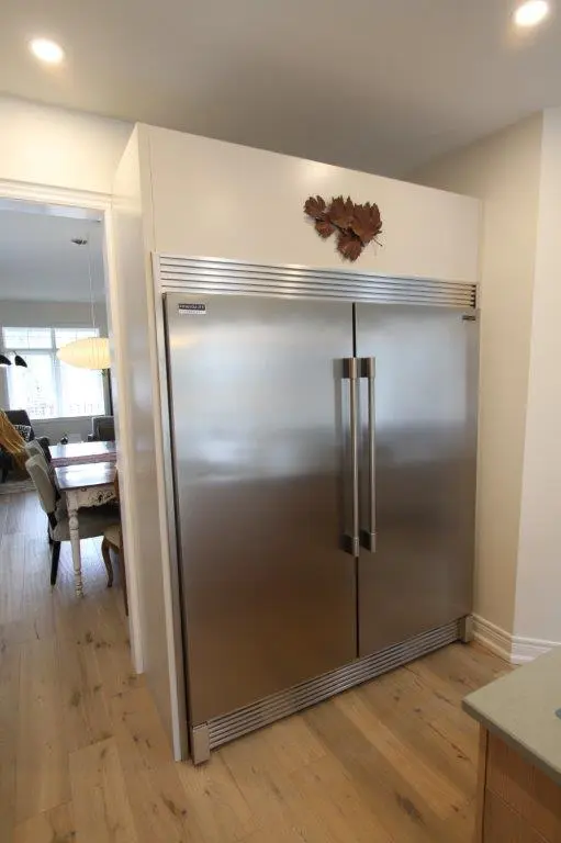 A large stainless steel refrigerator with double doors is built into a kitchen wall, with wooden flooring and a dining area visible in the background.
