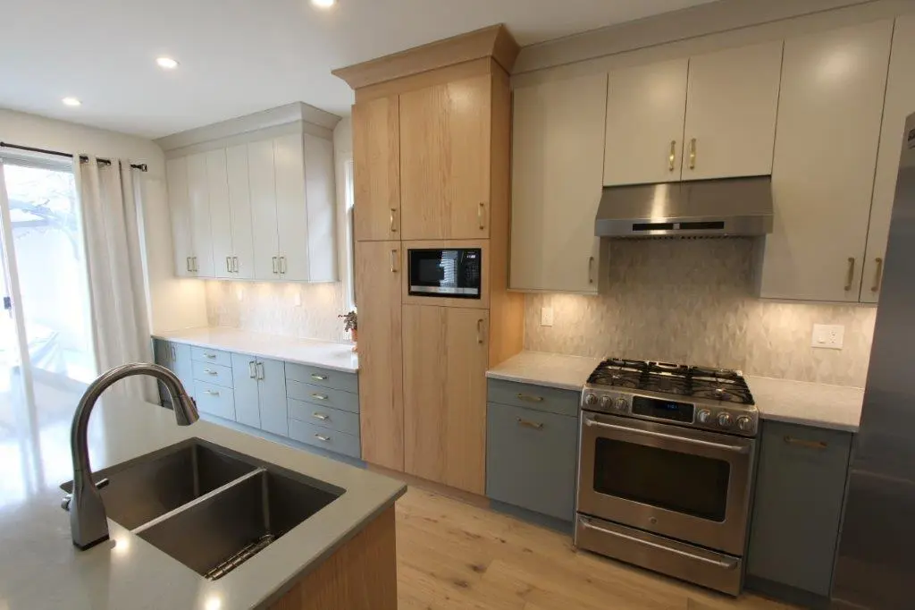 Modern kitchen interior with gray and white cabinetry, stainless steel appliances, a double sink with a sleek faucet, and a hood above the stove. There is also a microwave built into wooden cabinets and recessed lighting brightening the space.
