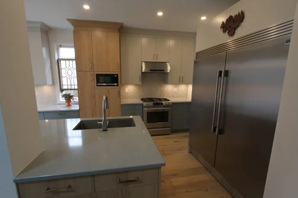 Modern kitchen with stainless steel appliances, light wood cabinets, gray quartz countertops, and a large metal refrigerator.
