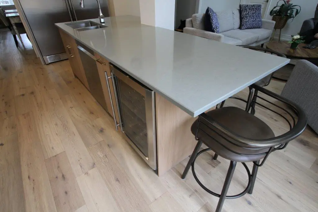 Modern kitchen with a large island featuring a light gray countertop, built-in wine fridge, and bar stool, set on a light wood floor.