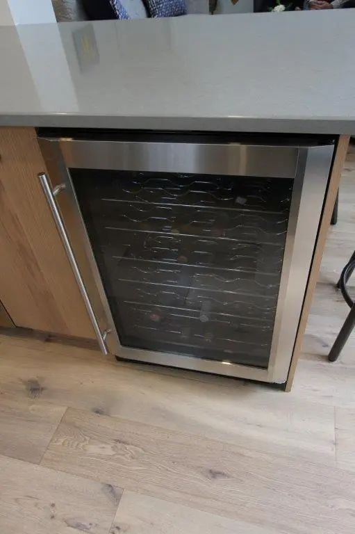 Under-counter wine refrigerator with a glass door, integrated into kitchen cabinetry on a light wooden floor.