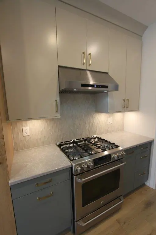 Modern kitchen with a stainless steel oven and range, light gray upper cabinets, teal lower cabinets, and a geometric tile backsplash.