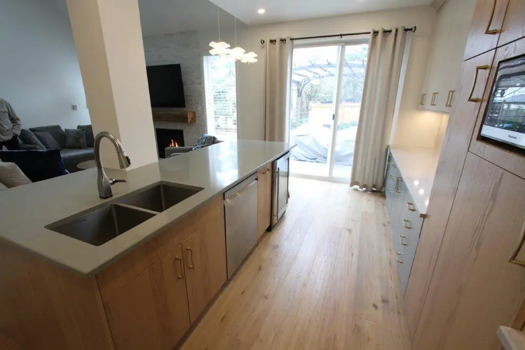 A modern kitchen with light wood flooring, featuring a large island with a double sink and dishwasher. There are built-in cabinets and a microwave on the right, and a living area with a fireplace and TV in the background, visible through a large glass door that opens to a patio.