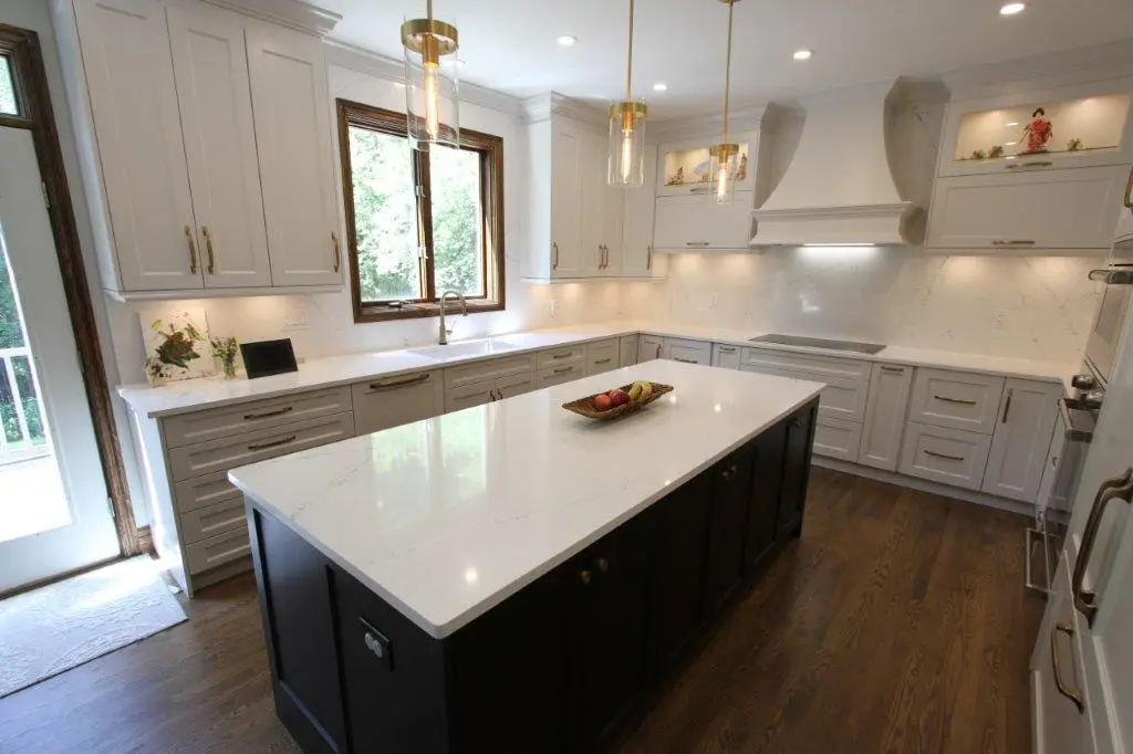 A modern kitchen featuring light-colored cabinets, a large central island with a dark base, quartz countertops, and wood flooring. The room is well-lit with pendant lights and recessed ceiling lights, and there are decorative shelves above the stove area.