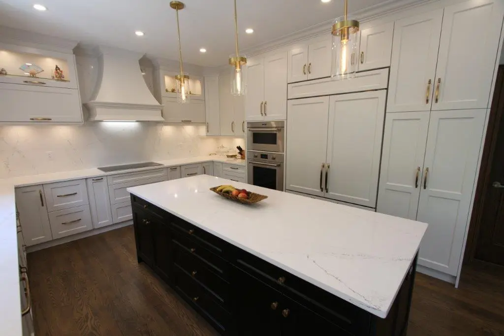 Modern kitchen with white cabinetry, a large quartz island, stainless steel appliances, and wooden flooring; pendant lighting hangs from the ceiling.