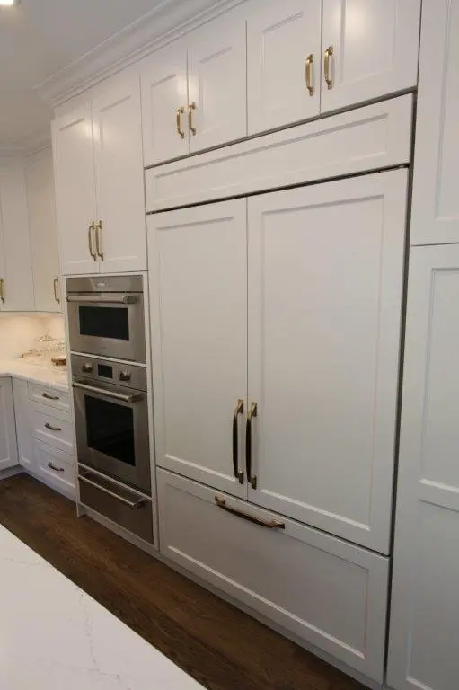 A modern kitchen featuring a built-in double oven next to a large refrigerator with panel doors, both surrounded by white cabinetry with gold handles.