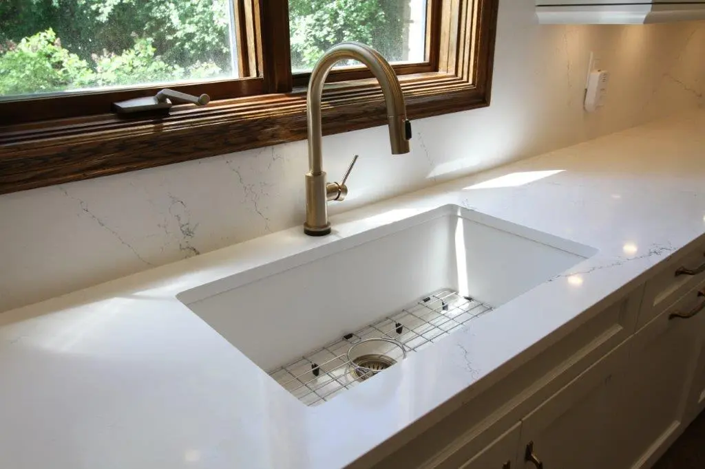 A modern kitchen sink with a sleek brass faucet set in a white quartz countertop, illuminated by natural light from a window above.
