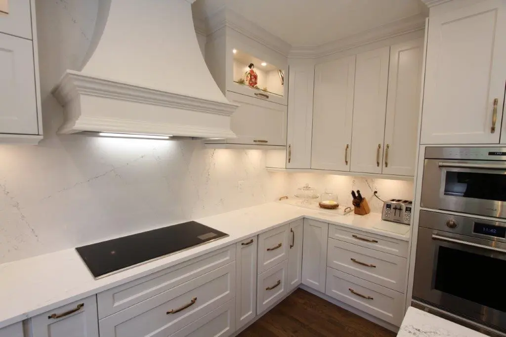 A modern kitchen with white cabinetry, featuring a sleek electric cooktop, stainless steel oven, and a toaster on the counter. The kitchen has under-cabinet lighting, a decorative range hood, and wooden flooring.