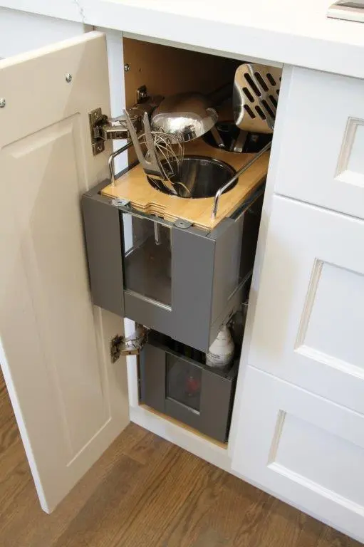 A slide out kitchen cabinet storage solution with grey compartments, containing various kitchen utensils and bottles, accessed through an open white cabinet door.