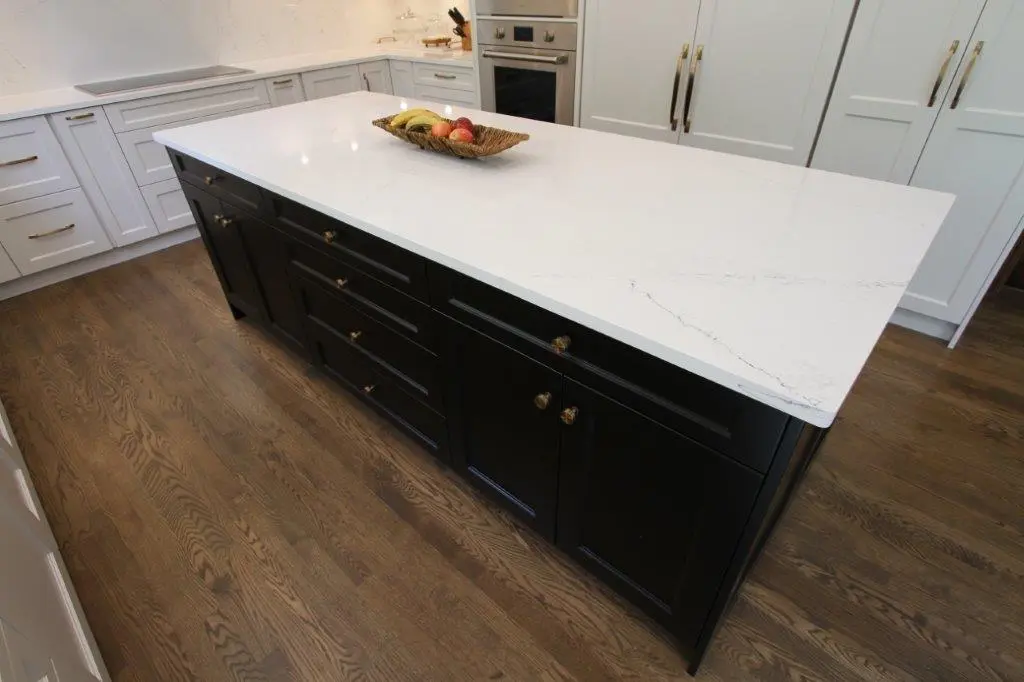 A modern kitchen with a large island featuring a white quartz countertop and dark cabinetry. The island is topped with a decorative basket containing assorted fruits. The kitchen includes white cabinets, brass handles, and a wooden floor.