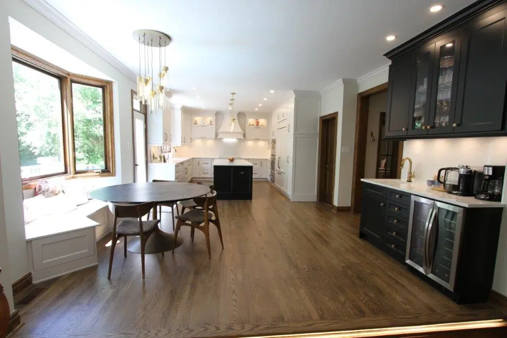 A modern kitchen with wooden flooring, white cabinets, and a central island with a black base. There's a large bay window seat, a round dining table with chairs, and a black cabinet with a built-in wine cooler and coffee maker. Pendant lighting hangs from the ceiling.