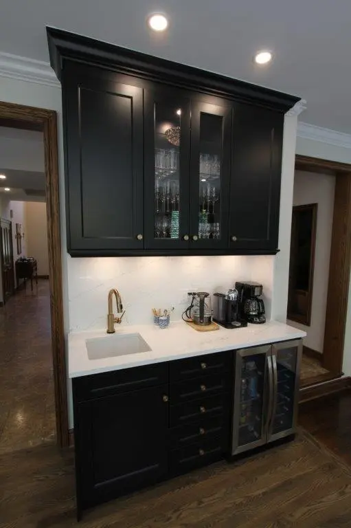 A stylish home bar setup with dark cabinetry, a white countertop, a small sink with a bronze faucet, and a mini fridge with a glass door. There are beverage appliances such as a coffee maker and espresso machine on the counter. Above, glassware is displayed in a cabinet with transparent doors.