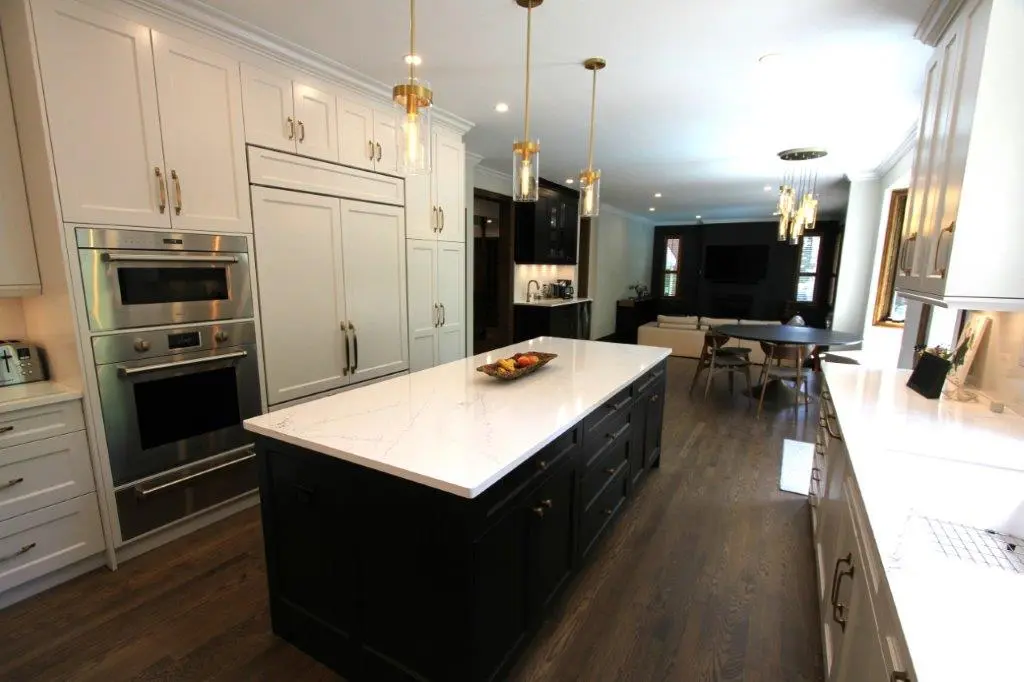 A modern kitchen with a large central island featuring a white quartz countertop and dark cabinetry. The kitchen includes built-in stainless steel appliances, white cabinetry, and pendant lighting. Hardwood floors extend into an adjacent dining and living area.