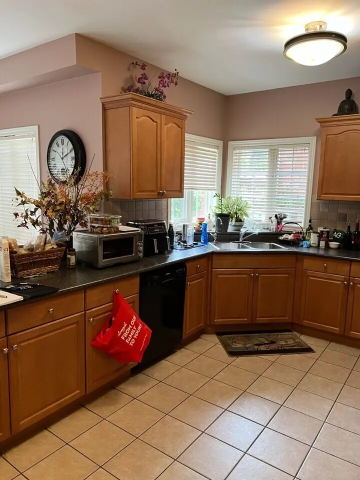 A before renovation kitchen with wooden cabinets, a tile floor, and ample natural light from large windows. The countertops are cluttered with various items. A decorative wall clock and artificial flowers add to the decor.