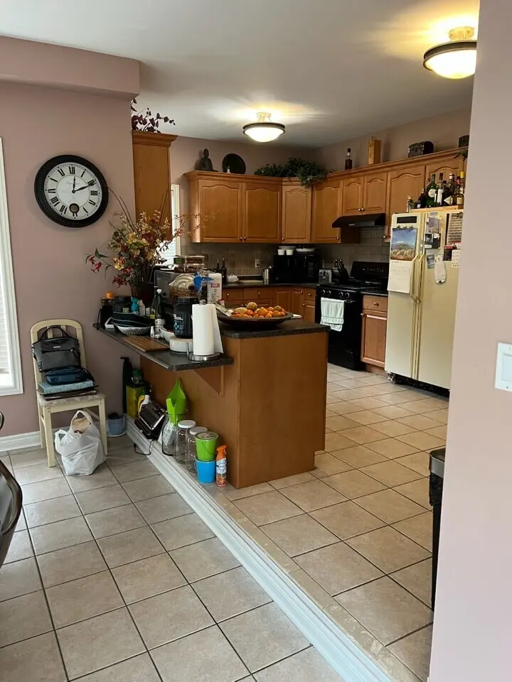 A before renovation kitchen with wooden cabinets, a refrigerator with magnets, a countertop with various items including a bowl of fruits, and a wall clock.