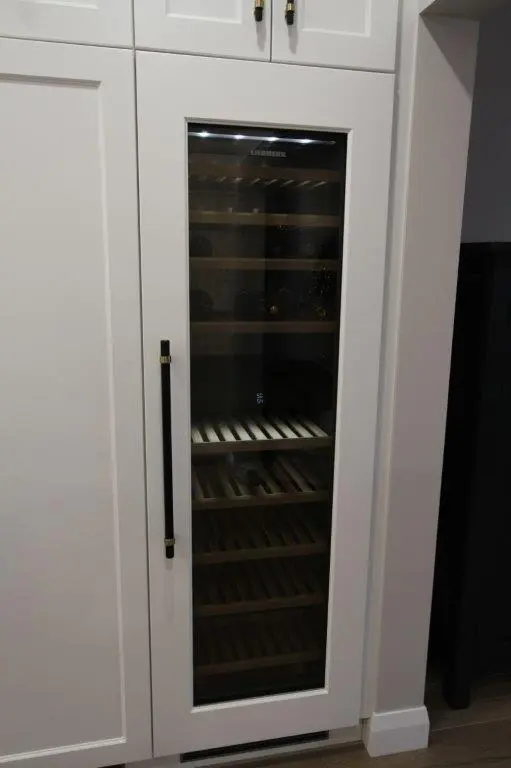 A built-in wine cooler with a glass door and several shelves for bottle storage, positioned within a white kitchen cabinet unit.