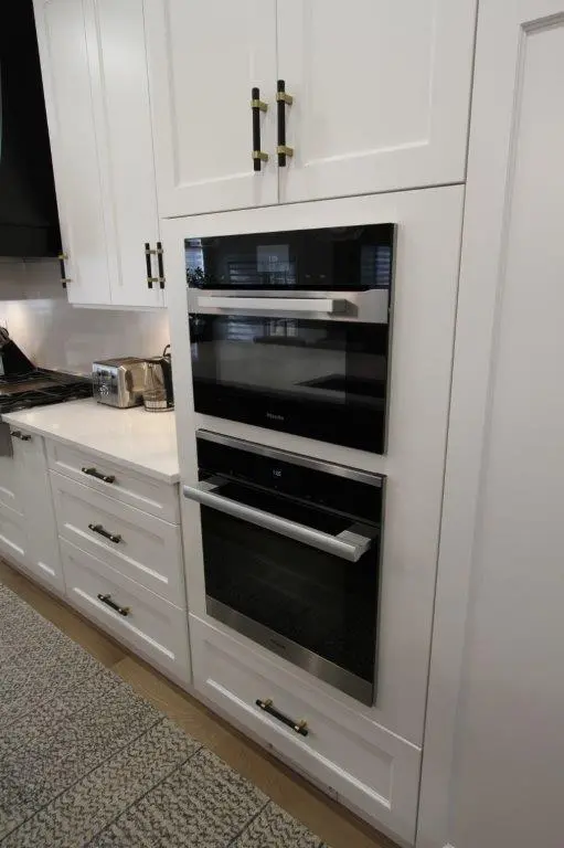 A modern kitchen with white cabinetry featuring an integrated double oven with stainless steel accents, a countertop with a toaster, and sleek black and gold handles on drawers and cupboards.