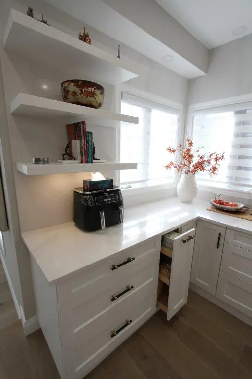 A modern kitchen corner with white cabinetry and countertops, featuring floating shelves above with decorative items. A vase with decorative foliage is placed near the window. A pull-out drawer is slightly open.