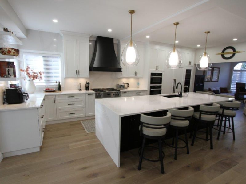 Modern kitchen with white cabinets, a large black range hood, a central island with white countertop and bar stools, and pendant lights overhead.