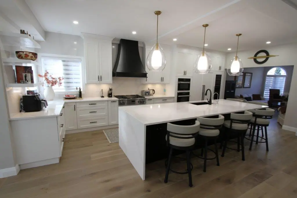Modern kitchen with white cabinets, a large black range hood, a central island with white countertop and bar stools, and pendant lights overhead.