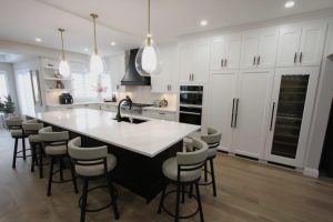 Modern kitchen featuring a large island with white countertop surrounded by bar stools, pendant lighting overhead, white cabinetry, an oven, and a built-in wine cooler in the background.
