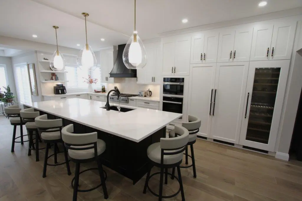 Modern kitchen featuring a large island with white countertop surrounded by bar stools, pendant lighting overhead, white cabinetry, an oven, and a built-in wine cooler in the background.