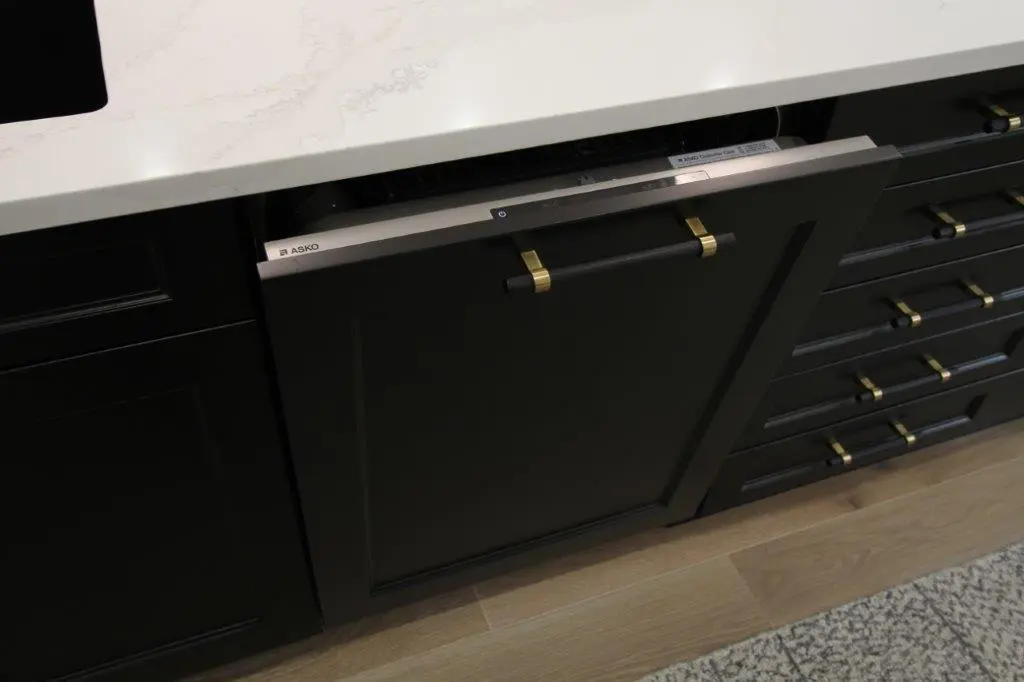 A kitchen with black cabinetry featuring brass handles and a partially open built-in dishwasher beneath a white countertop.