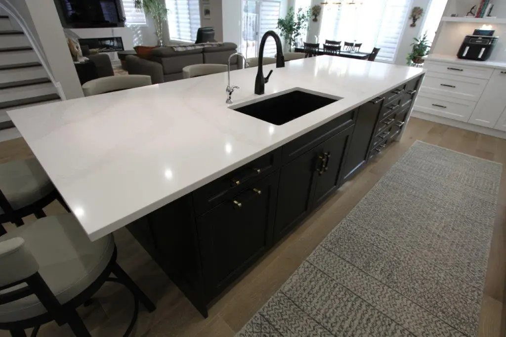 A modern kitchen featuring a large white quartz countertop island with a black sink and faucet. The island has dark cabinetry with gold handles and is set on a light wooden floor with a patterned runner rug. In the background, there is a seating area with a dark sectional sofa, dining table, and windows with blinds.