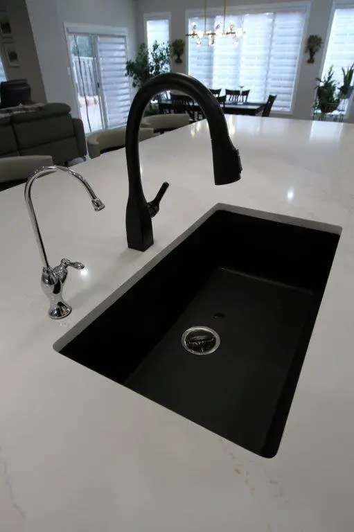 A modern kitchen sink with a black faucet and a separate smaller chrome faucet on a white countertop, set in a spacious kitchen with large windows in the background.