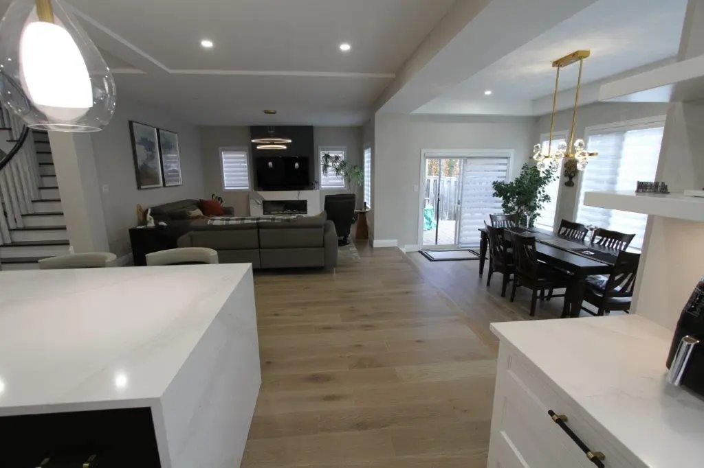 Open-plan living room and dining area with wooden flooring, featuring a large gray sofa facing a wall-mounted TV, a dining table with chairs near sliding glass doors, and a white quartz kitchen island in the foreground.