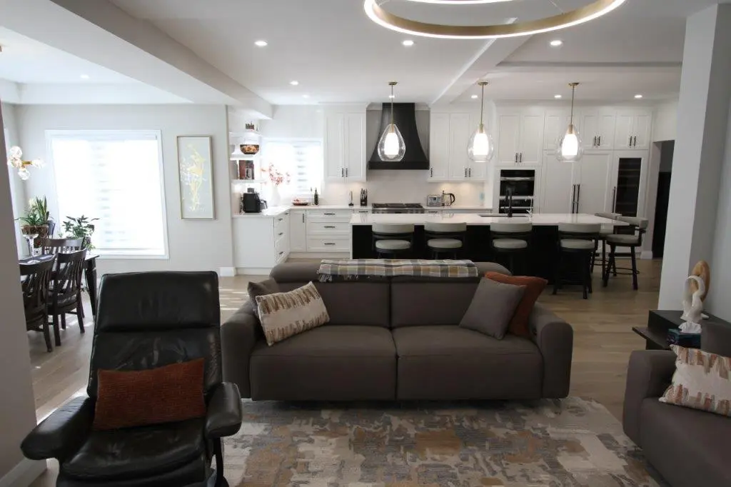 Open-concept living room and kitchen with modern furniture, including a grey sofa and armchair, a large kitchen island with pendant lights, and a dining table by a window.