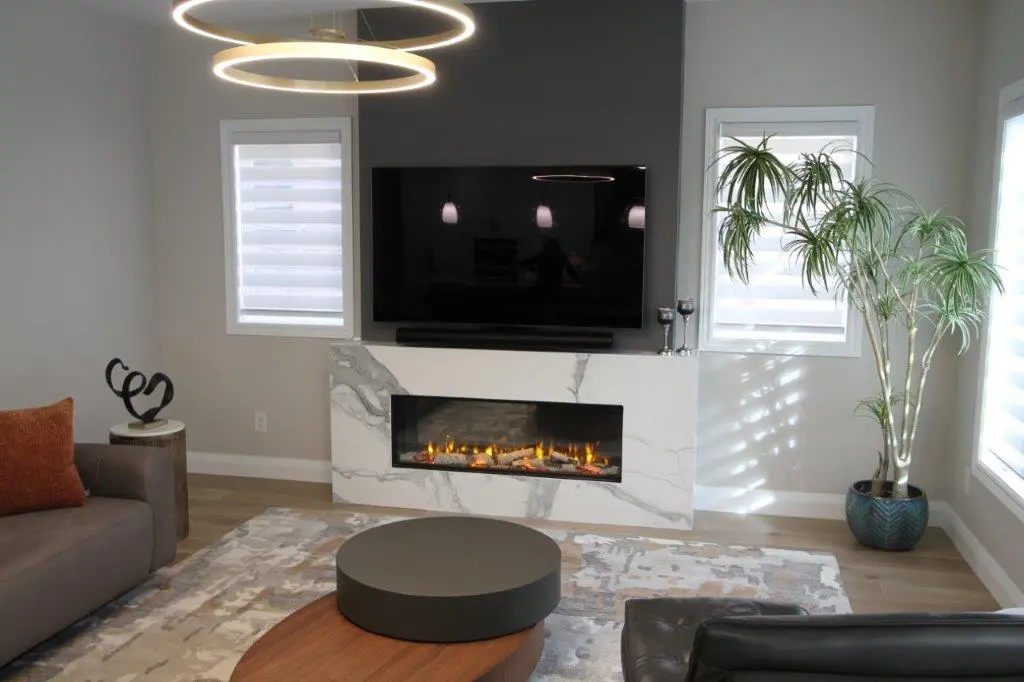 Modern living room with a large flat-screen TV above a quartz fireplace, circular pendant lights, a gray sofa, a round coffee table, and a potted plant by the window.