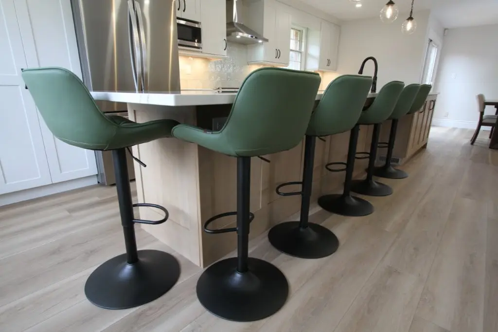 A modern kitchen with a wooden floor and six green swivel bar stools at a light wood kitchen island. Stainless steel appliances and pendant lights are visible in the background.