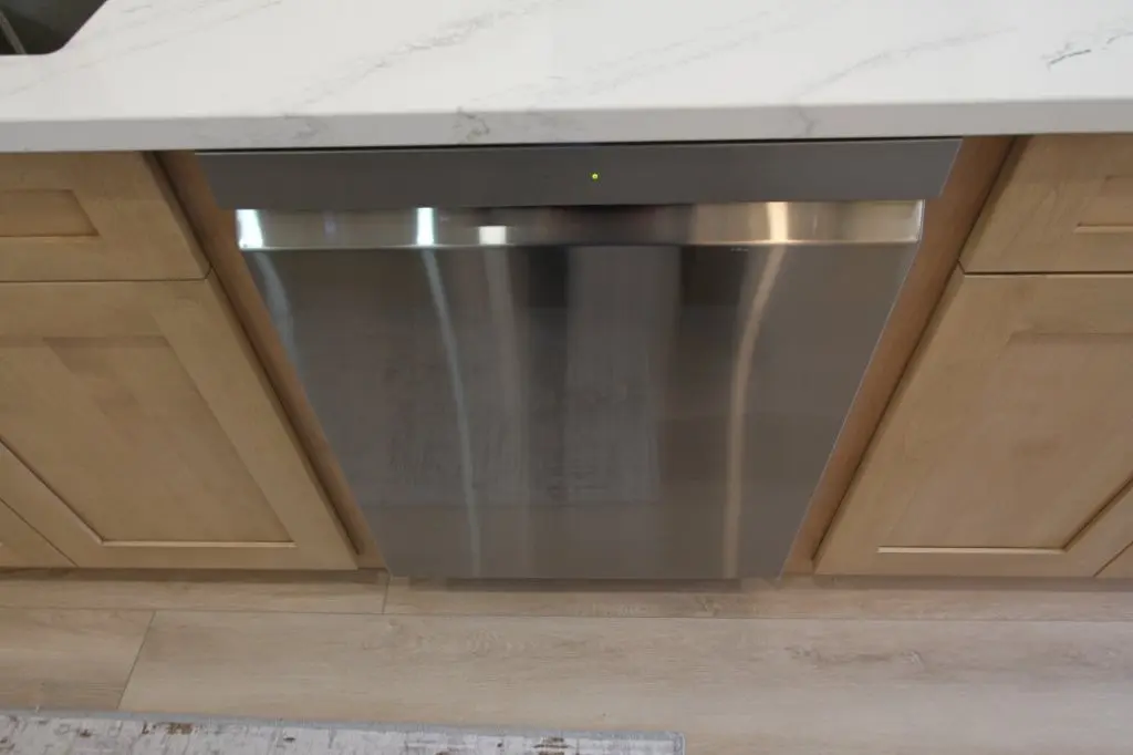 Stainless steel dishwasher integrated into a kitchen with light wood cabinets and a quartz countertop.