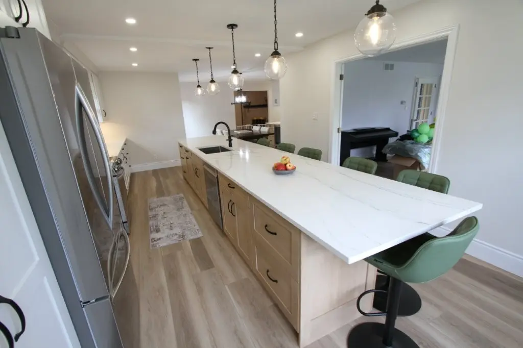Modern kitchen with a long white marble island countertop, equipped with a sink, hanging pendant lights, green barstools, and a stainless steel refrigerator on the side.
