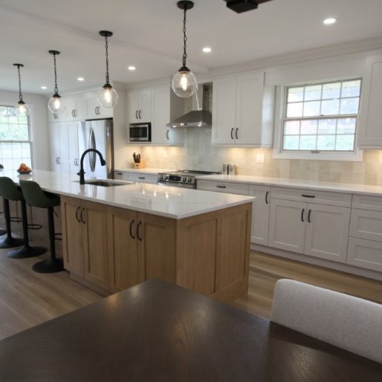 A modern kitchen with white cabinets, stainless steel appliances, and a large island with barstools. The island features a wooden base and a white countertop with a built-in sink and faucet. Pendant lights hang above the island, and there is a window in the background letting in natural light.