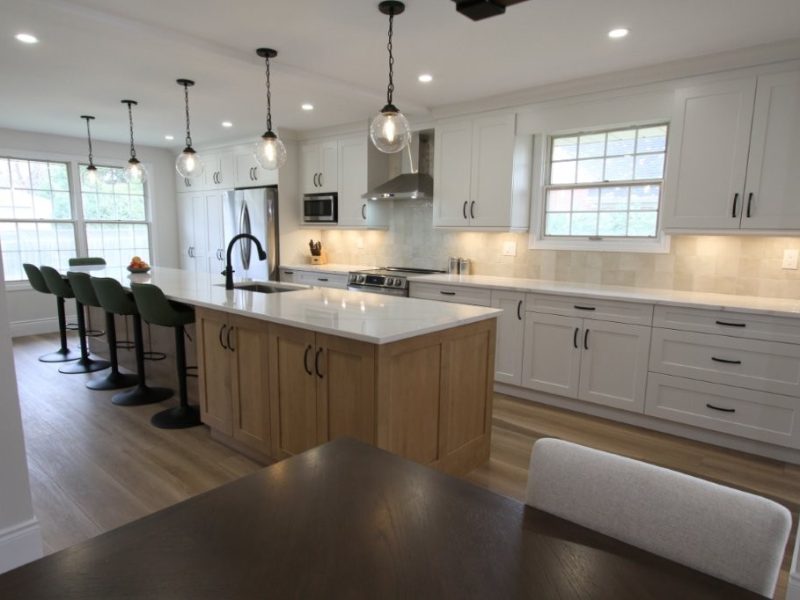 A modern kitchen with white cabinets, stainless steel appliances, and a large island with barstools. The island features a wooden base and a white countertop with a built-in sink and faucet. Pendant lights hang above the island, and there is a window in the background letting in natural light.